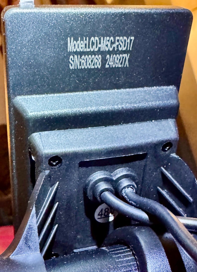 May include: Close-up of a black electronic component with the text "Model: LCD-M5C-FSD17" and "S/N:608268 240927X" printed in white. Two black cables are connected to the component. A sticker with the number "48" is visible.