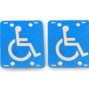 May include: Two blue, square handicap signs with a white wheelchair symbol. Each sign has a textured surface and several small holes for mounting. The signs are designed to indicate accessibility.