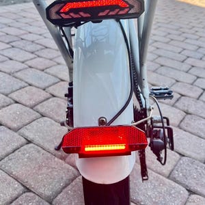May include: A white electric bicycle with a rear rack and two red taillights. The bicycle has a black tire and a black fender.