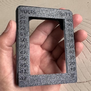 May include: A textured, dark gray rectangular gauge with the words "VOLTS" and "BATT" printed on it. The gauge has a series of numbers from 0 to 100, and 42 to 53. The gauge is held in a person's hand.