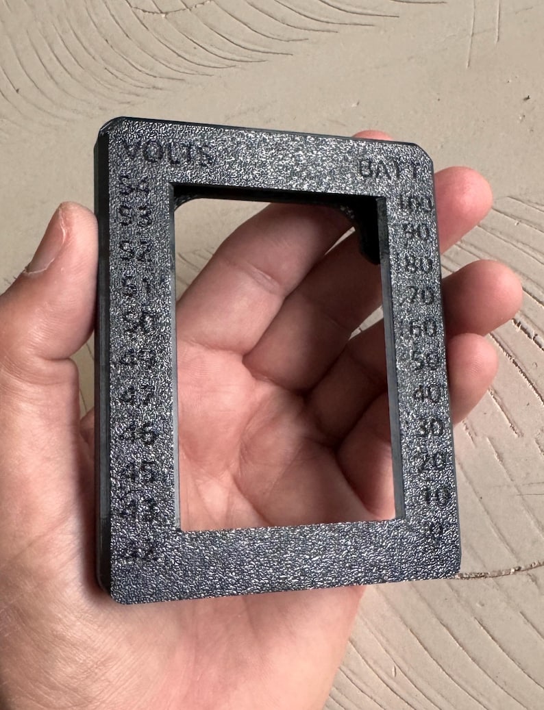 May include: A textured, dark gray rectangular gauge with the words "VOLTS" and "BATT" printed on it. The gauge has a series of numbers from 0 to 100, and 42 to 53. The gauge is held in a person's hand.