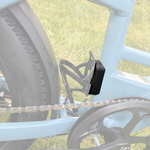 May include: A black bicycle light is mounted on a light blue bicycle frame, next to a black water bottle cage. The bicycle tire and chain are also visible. The background is a blurred green.