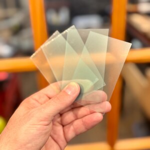 May include: A hand holding a small stack of translucent, square-shaped plastic sheets. The sheets have a slightly frosted appearance and are overlapping. The background is blurred, with hints of orange and brown.