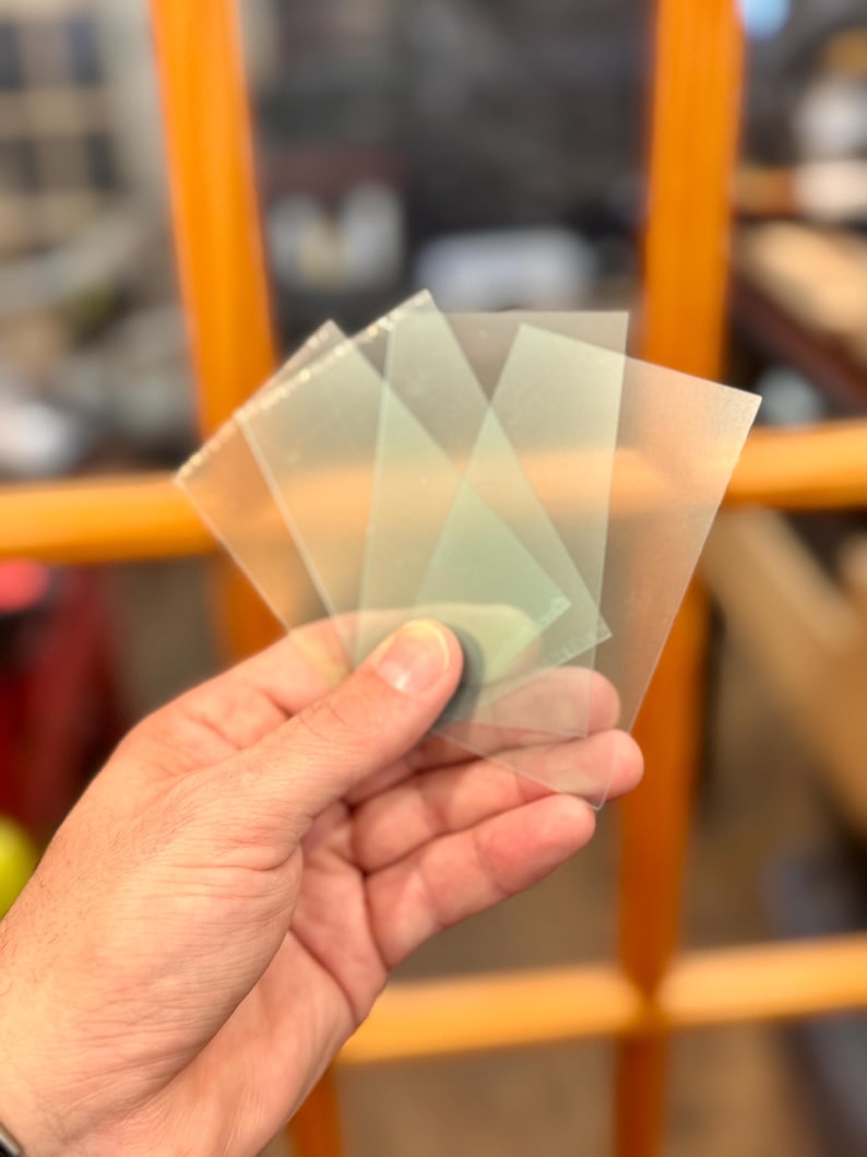 May include: A hand holding a small stack of translucent, square-shaped plastic sheets. The sheets have a slightly frosted appearance and are overlapping. The background is blurred, with hints of orange and brown.