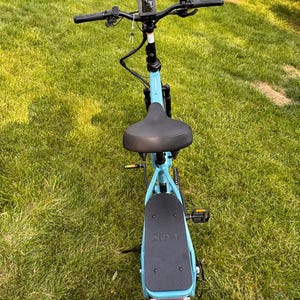 May include: A light blue electric scooter with a black seat, handlebars, and foot platform. The scooter has a digital display, a rear light, and a black tire. The scooter is on a grassy surface.
