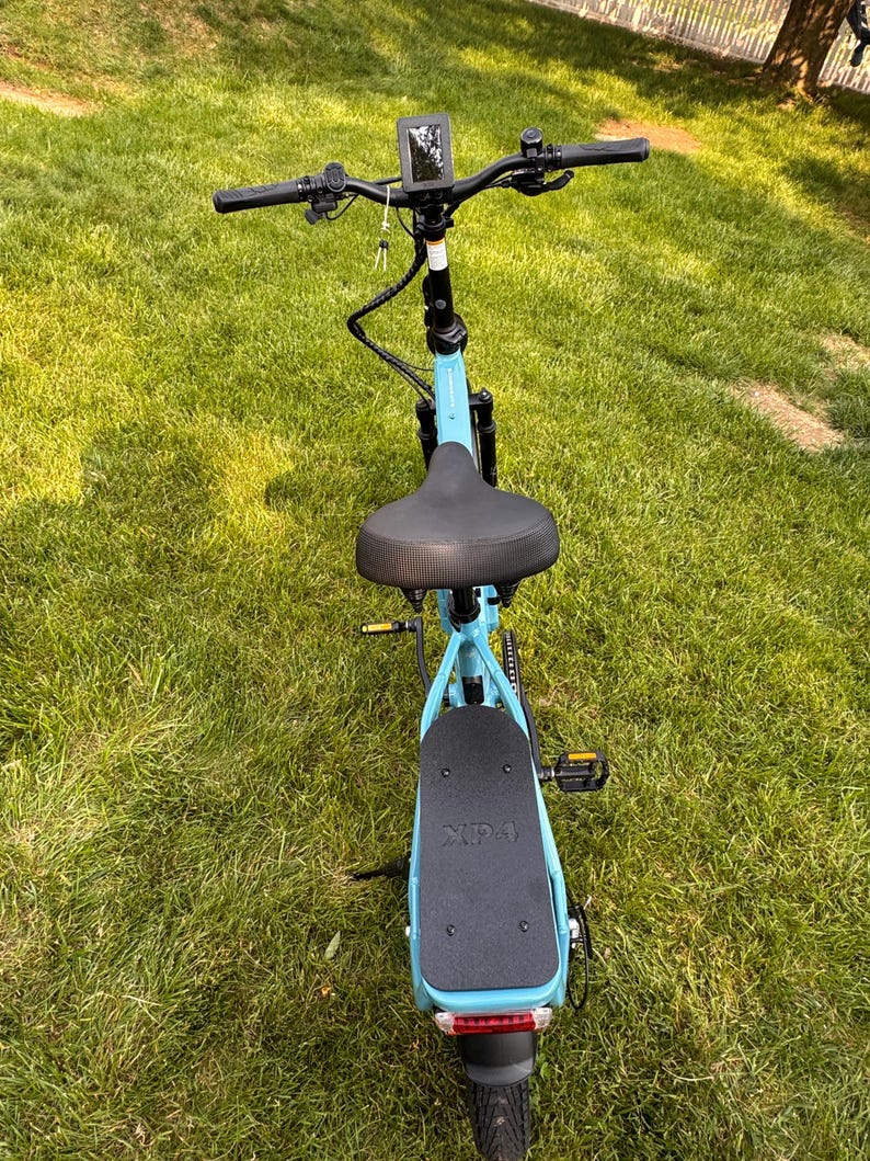 May include: A light blue electric scooter with a black seat, handlebars, and foot platform. The scooter has a digital display, a rear light, and a black tire. The scooter is on a grassy surface.