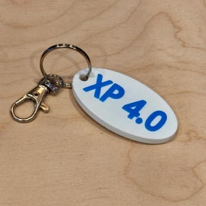 May include: White oval keychain with the text "XP 4.0" in blue. The keychain has a silver metal ring and clasp. The background is a light brown wooden surface.