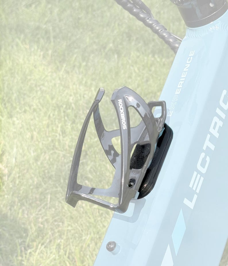May include: A gray ROCKBROS water bottle cage mounted on a light blue bicycle frame. The cage is made of a lightweight material and has a black, padded section. The bicycle frame has the word "LECTRIC" printed on it.