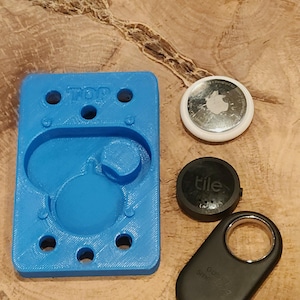 May include: A blue 3D printed holder with six holes and the word "TOP" printed on it. There are three black and silver Tile trackers next to the holder.