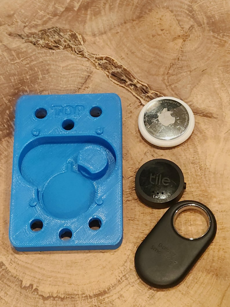 May include: A blue 3D printed holder with six holes and the word "TOP" printed on it. There are three black and silver Tile trackers next to the holder.