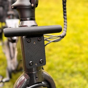 May include: Close-up of a black electric scooter's handlebar and stem. The handlebar is a straight, cylindrical bar. The stem features a textured black plate secured with four screws. A black cable extends from the stem.