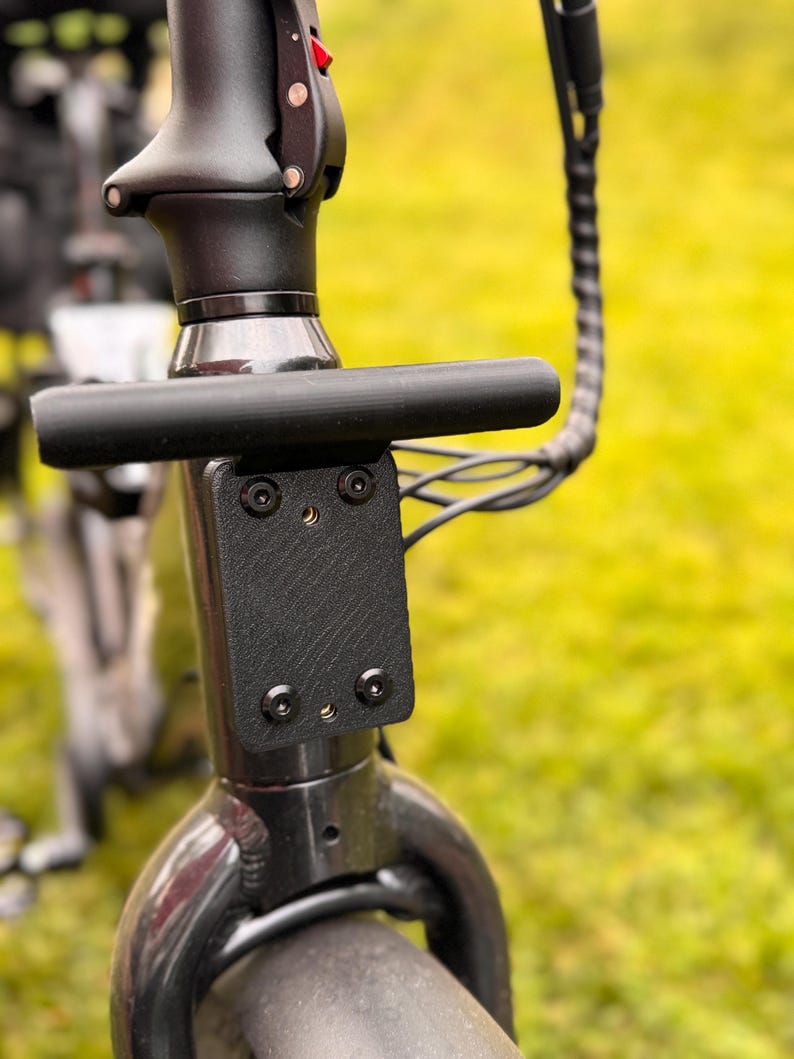 May include: Close-up of a black electric scooter's handlebar and stem. The handlebar is a straight, cylindrical bar. The stem features a textured black plate secured with four screws. A black cable extends from the stem.