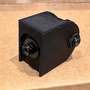 May include: A black, 3D-printed, angular object with two black bolts. The object has a textured surface and a rectangular shape with rounded edges. The bolts are mounted on circular black plates.