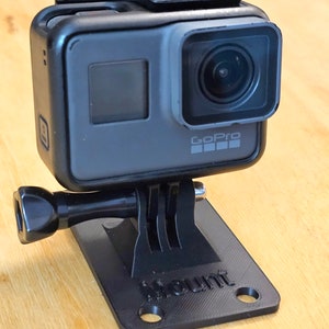 May include: A black GoPro camera mounted on a black plastic base with the word "Mount" printed on it. The camera has a black lens and a small screen on the back.