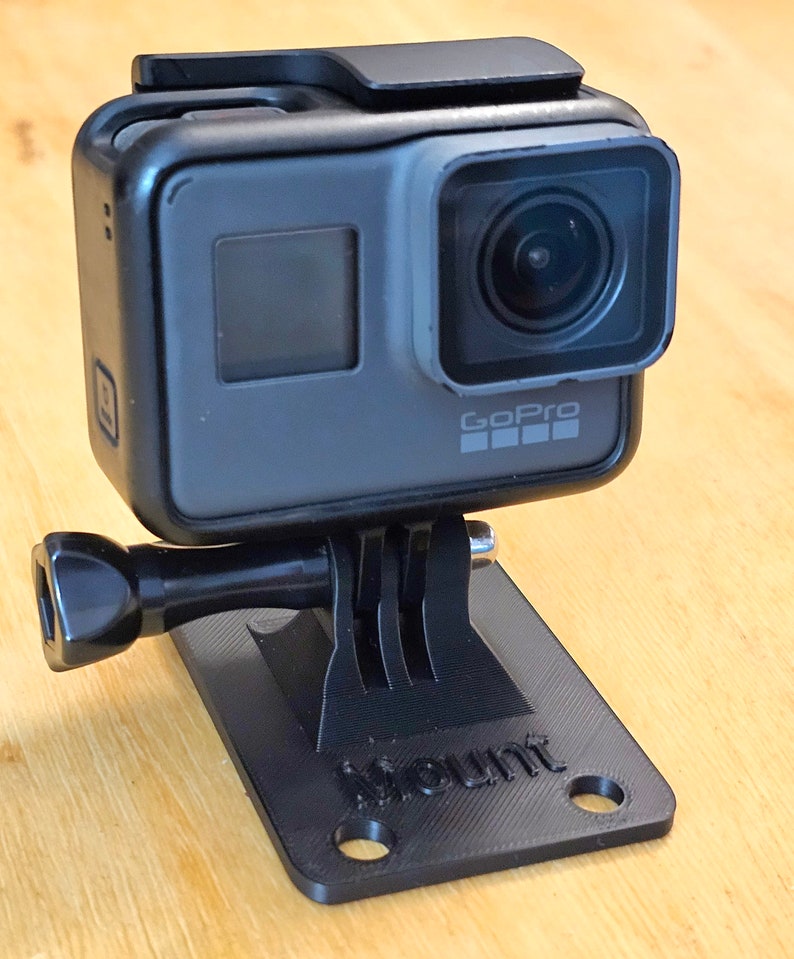 May include: A black GoPro camera mounted on a black plastic base with the word "Mount" printed on it. The camera has a black lens and a small screen on the back.
