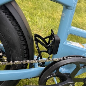 May include: Close-up of a light blue bicycle frame with a black water bottle cage. The cage is mounted on the frame near the chain and tire. The tire is black and has a thick tread. The chain and gears are also visible.