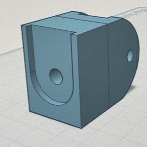 May include: A light blue, 3D-rendered bracket with a U-shaped cutout and two circular holes. The bracket has a rectangular base and a rounded side. The object is set against a grid background.
