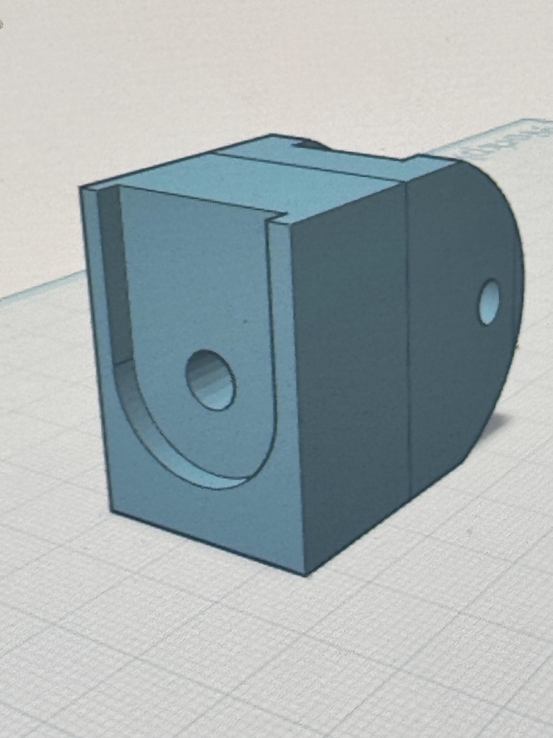 May include: A light blue, 3D-rendered bracket with a U-shaped cutout and two circular holes. The bracket has a rectangular base and a rounded side. The object is set against a grid background.