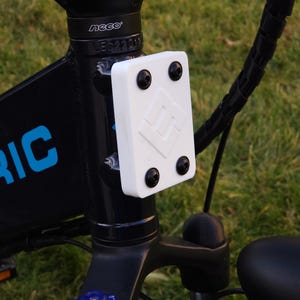 May include: Close-up of a black electric bicycle with a white plastic cover with four black screws. The cover has a logo with the letter "M" inside a diamond shape. The bicycle has a black handlebar and a black seat.