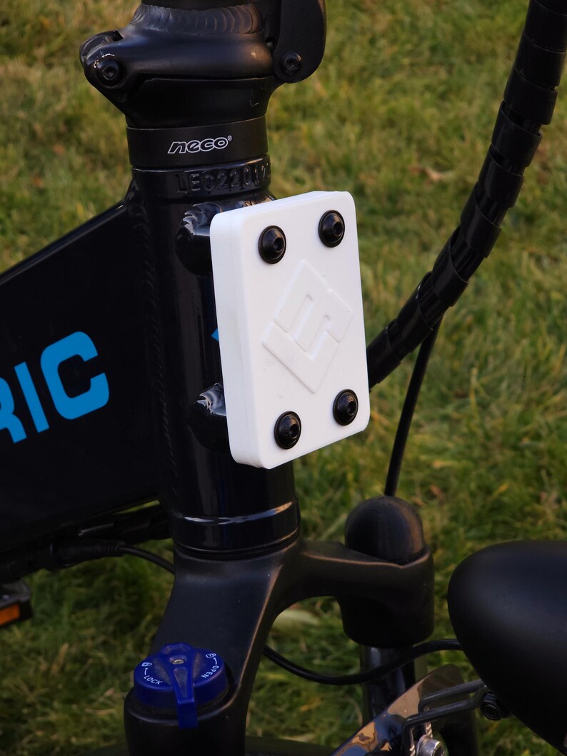 May include: Close-up of a black electric bicycle with a white plastic cover with four black screws. The cover has a logo with the letter "M" inside a diamond shape. The bicycle has a black handlebar and a black seat.