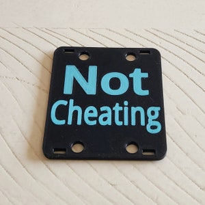 May include: Black square sign with rounded corners and four holes. The sign says "Not Cheating" in light blue text.