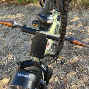 May include: Close-up of an electric bicycle's handlebars, featuring a black frame and a headlight with the word "Lectric" printed on it. The handlebars have orange turn signals on each end. The bike is green.