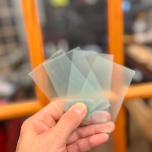 May include: A hand holding several translucent, square-shaped, light blue and clear plastic sheets. The sheets are slightly overlapping, with a few visible in the foreground. The background is blurred, with hints of orange and other colors.