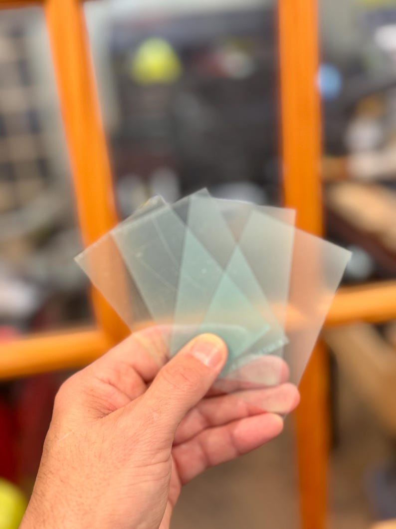 May include: A hand holding several translucent, square-shaped, light blue and clear plastic sheets. The sheets are slightly overlapping, with a few visible in the foreground. The background is blurred, with hints of orange and other colors.