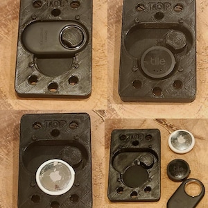 May include: Black 3D-printed holders for tracking devices. The holders are rectangular with rounded edges and feature the word "TOP" at the top. The holders are designed to fit various tracking devices, including Tile and Apple AirTag.