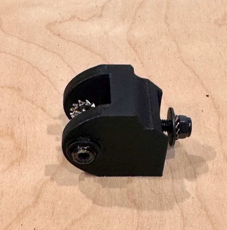 May include: A black metal clamp with a bolt and washer. The clamp has a curved design with a gear-like component visible inside. The object is set against a light brown wooden surface.