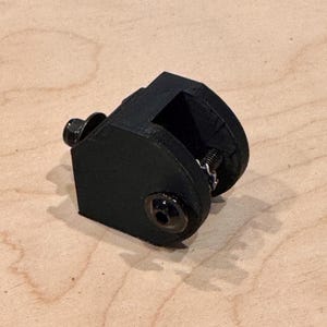 May include: A black, 3D-printed clamp with a spring and two screws. The clamp has a rectangular opening and a curved top. The screws and spring are silver. The clamp is sitting on a light brown wooden surface.
