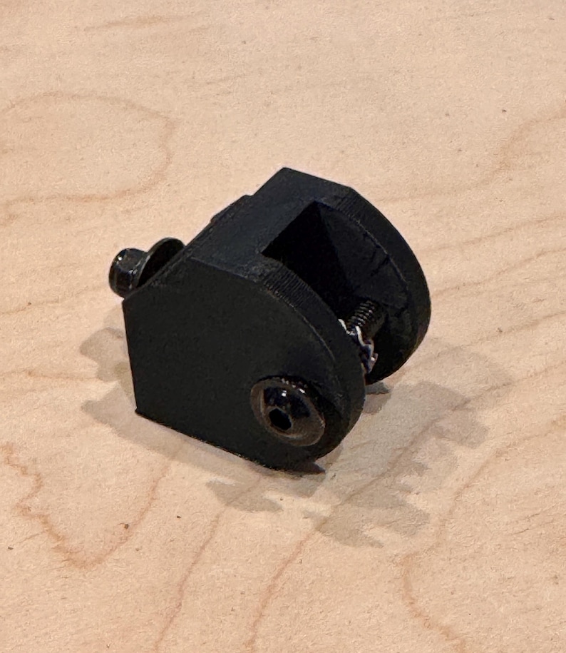 May include: A black, 3D-printed clamp with a spring and two screws. The clamp has a rectangular opening and a curved top. The screws and spring are silver. The clamp is sitting on a light brown wooden surface.