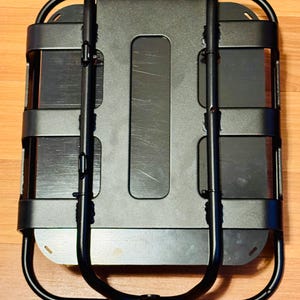 May include: A black metal cargo carrier with a rectangular base and a curved, U-shaped handle. The carrier has horizontal bars and a central rectangular opening. The design is utilitarian and functional, suitable for carrying items.