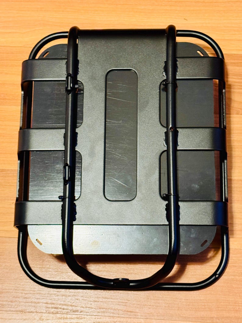 May include: A black metal cargo carrier with a rectangular base and a curved, U-shaped handle. The carrier has horizontal bars and a central rectangular opening. The design is utilitarian and functional, suitable for carrying items.
