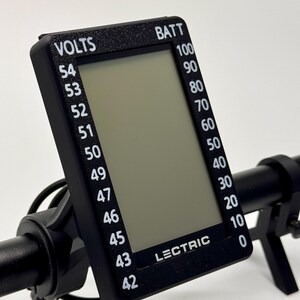 May include: A black digital display with the text "VOLTS" and "BATT" is mounted on a black handlebar. The display shows voltage and battery percentage readings. The brand name "LECTRIC" is printed on the display.