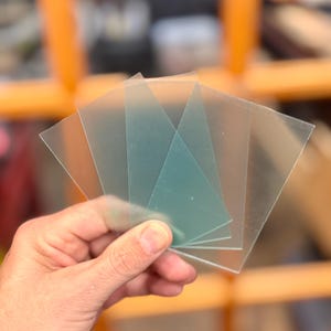 May include: Four clear plastic sheets, one of which is a light green color, are held in a hand. The sheets are rectangular and appear to be used for crafting or other projects.