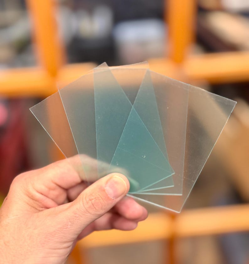 May include: Four clear plastic sheets, one of which is a light green color, are held in a hand. The sheets are rectangular and appear to be used for crafting or other projects.