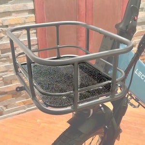 Puede incluir: Una cesta de bicicleta de metal negro fijada a la parte delantera de una bicicleta. La cesta tiene forma rectangular con un diseño de rejilla y una base negra texturizada. Se ven el neumático delantero y el guardabarros de la bicicleta.