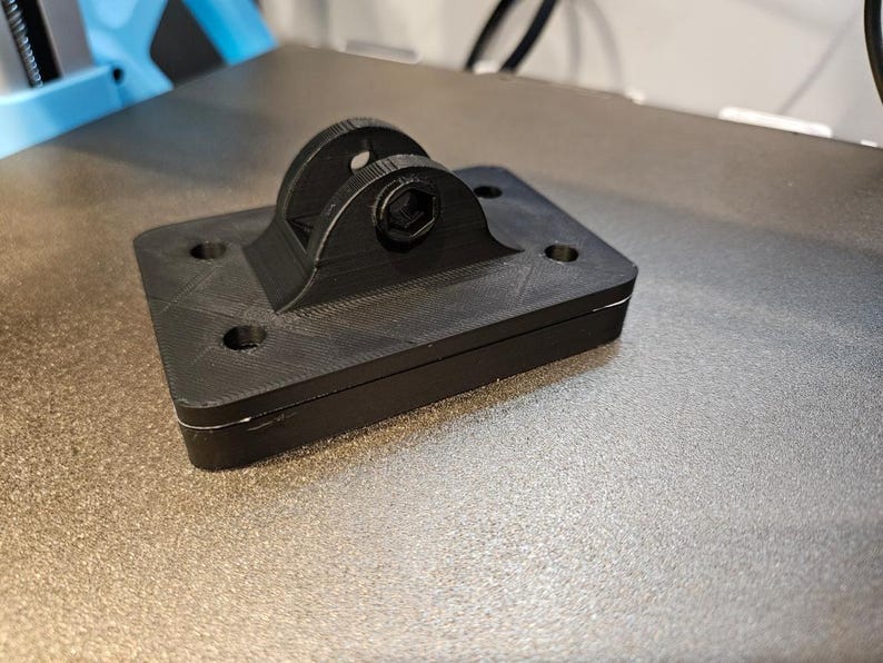 May include: A black, 3D-printed camera mount with a rectangular base and a curved top section. The mount has a textured surface and includes mounting holes and a hex screw for securing a camera. The item is designed for attaching a camera to a surface.