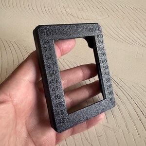 May include: A black rectangular tool with a textured surface, held in a hand. The tool has a square opening and is marked with numbers from 0 to 100, and the word "VOLTS".