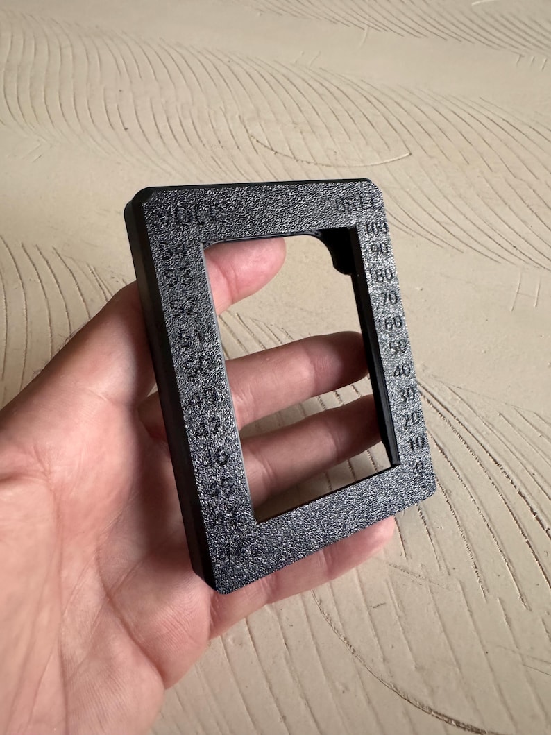May include: A black rectangular tool with a textured surface, held in a hand. The tool has a square opening and is marked with numbers from 0 to 100, and the word "VOLTS".