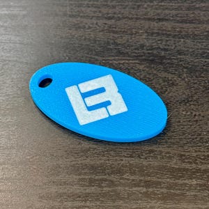 May include: A light blue, oval-shaped keychain with a white logo. The keychain has a hole at the top for attachment. The logo appears to be the letters "L" and "B" stacked on top of each other.