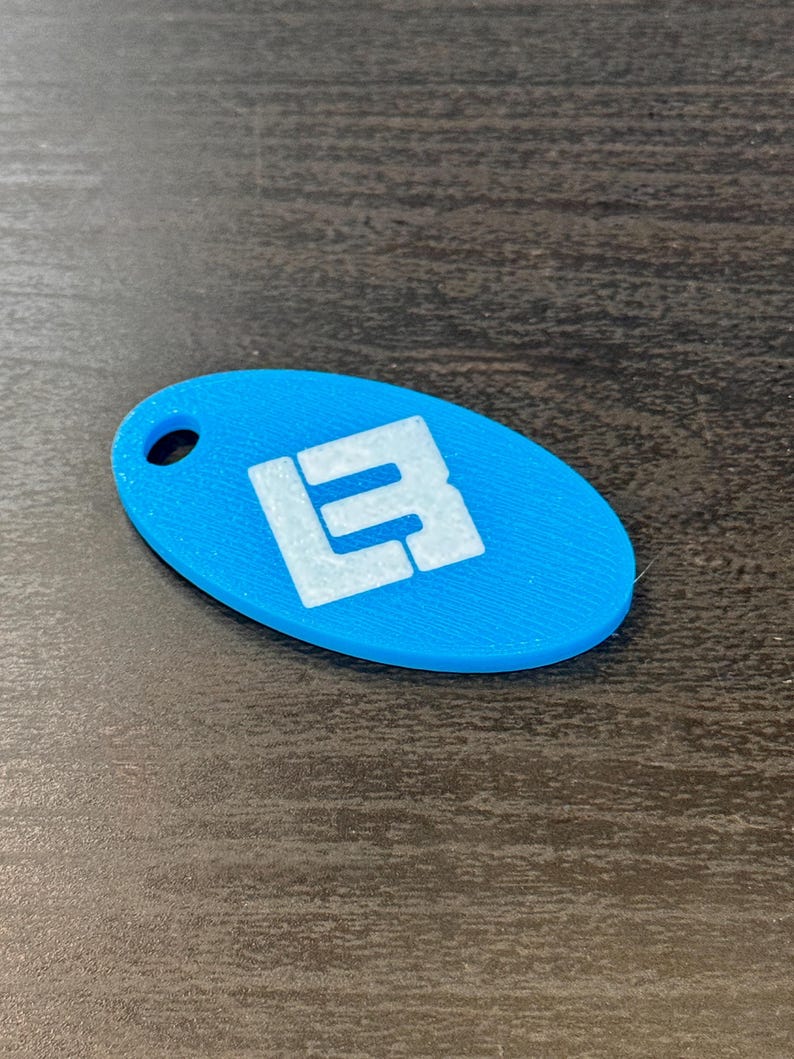 May include: A light blue, oval-shaped keychain with a white logo. The keychain has a hole at the top for attachment. The logo appears to be the letters "L" and "B" stacked on top of each other.