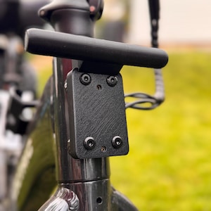 May include: Close-up of a black bicycle handlebar with a textured mounting plate. The handlebar is cylindrical and mounted on a black frame with visible bolts. The background is blurred green grass.