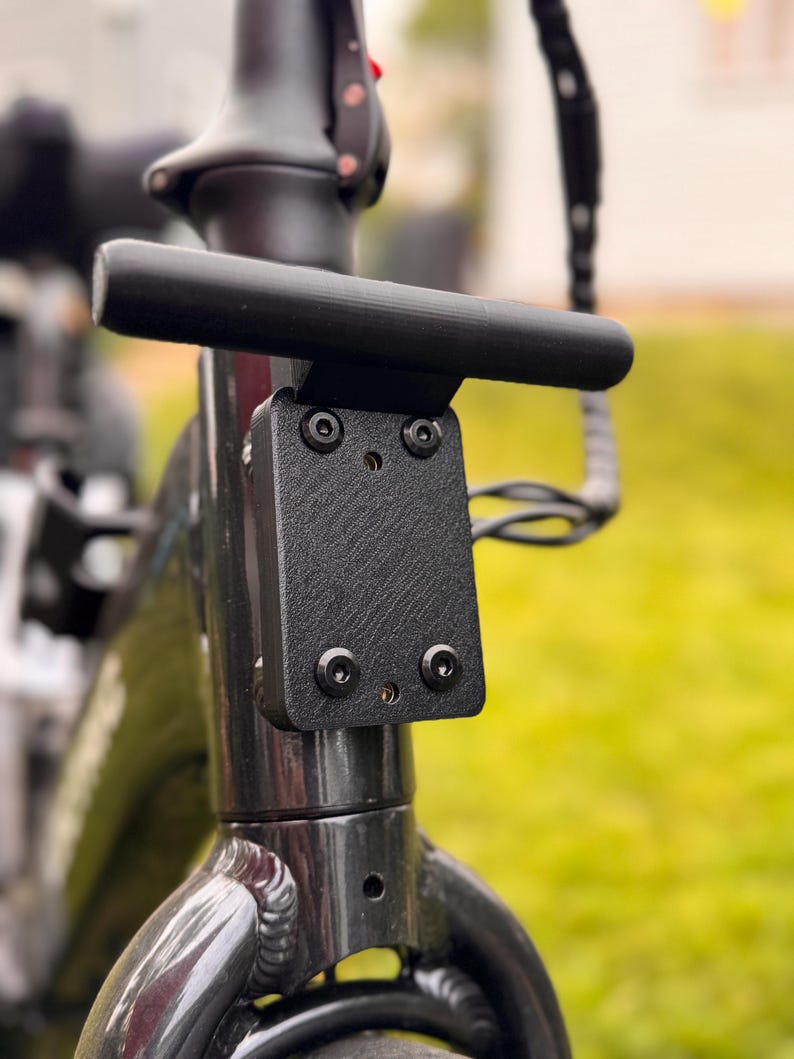 May include: Close-up of a black bicycle handlebar with a textured mounting plate. The handlebar is cylindrical and mounted on a black frame with visible bolts. The background is blurred green grass.