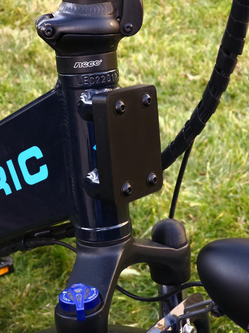 May include: Close-up of a black electric bicycle with a black metal plate attached to the frame. The plate has several screws and the text "neco" is visible on the frame.