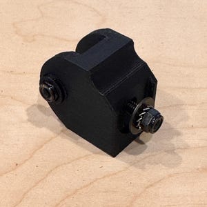 May include: A black, 3D-printed object with a rectangular base and a curved top. Two bolts and washers are attached to the sides. The object appears to be a mechanical component or part.