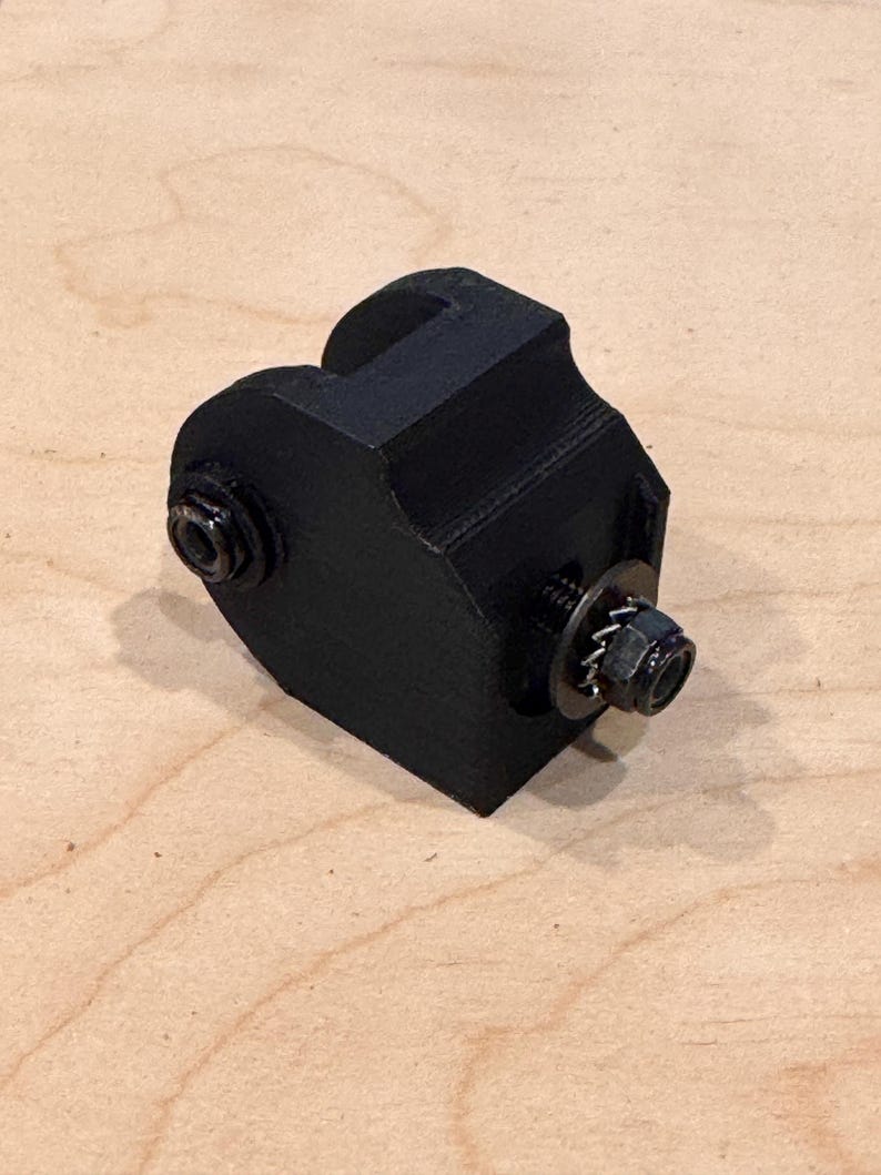 May include: A black, 3D-printed object with a rectangular base and a curved top. Two bolts and washers are attached to the sides. The object appears to be a mechanical component or part.