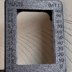 May include: A textured black rectangular frame with the words "VOLTS" and "BATT" printed on it. The frame has a scale from 0 to 100, with numbers and markings. The frame is made of a dark material.