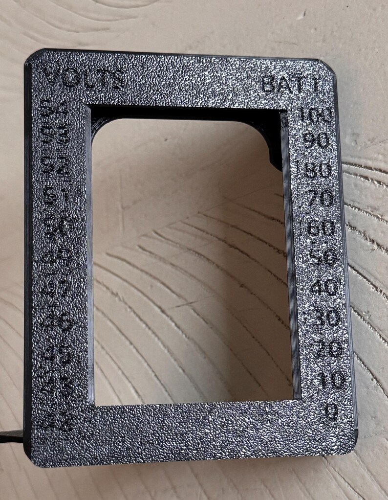 May include: A textured black rectangular frame with the words "VOLTS" and "BATT" printed on it. The frame has a scale from 0 to 100, with numbers and markings. The frame is made of a dark material.